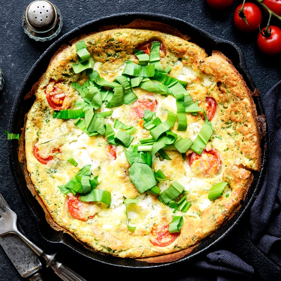 A frittata with tomatoes and green herbs in a cast iron skillet, with cherry tomatoes and cutlery.